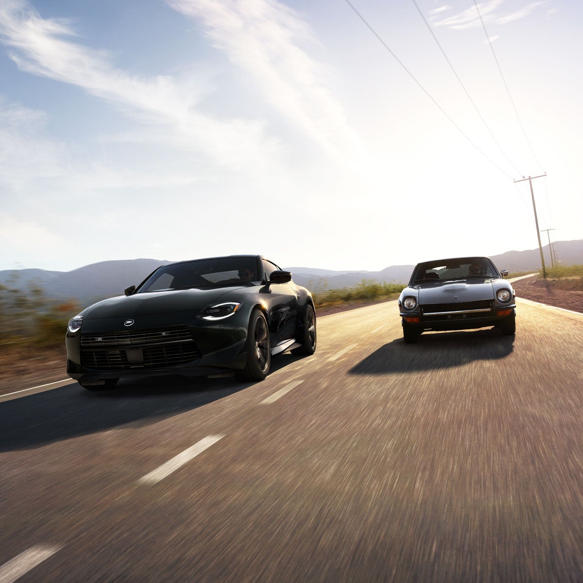 Two black Nissan Z sports cars, one modern and one classic, driving side-by-side on a highway at sunset.